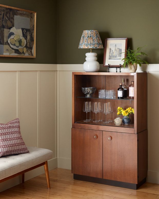 A warmly styled interior corner featuring a mid-century timber drinks cabinet with glassware and bottles, positioned against dark olive-green walls with cream-painted vertical wall panelling. A white ceramic lamp, framed artwork, and potted fern sit on top of the cabinet. Part of a light upholstered bench with a patterned cushion is visible at left, and the room has a timber floor.