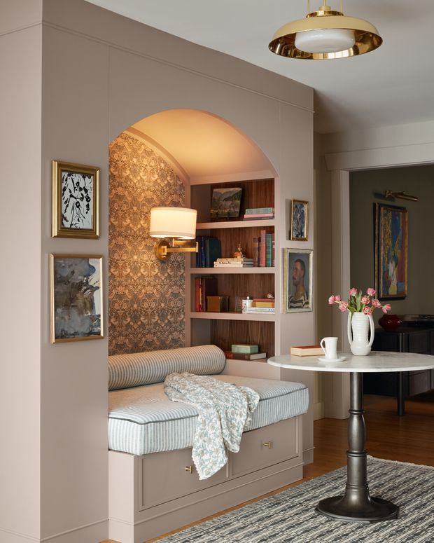 A refined traditional interior featuring a built-in upholstered window seat with drawers, framed by an arched alcove lined with ornate patterned wallpaper. Integrated dark timber shelving displays books and artwork, while a round marble-topped pedestal table and black piano occupy the adjoining sitting area. Taupe wall panelling, framed art, a striped rug, floral arrangement, and a brass pendant create a warm, elegant and layered atmosphere.
