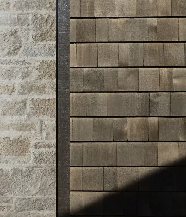 Close-up architectural detail showing a pale rough masonry wall beside a dark metal trim and a facade clad in staggered horizontal rows of narrow brown timber-like shingles or boards. Strong sunlight and shadow emphasize the layered texture and material contrast.