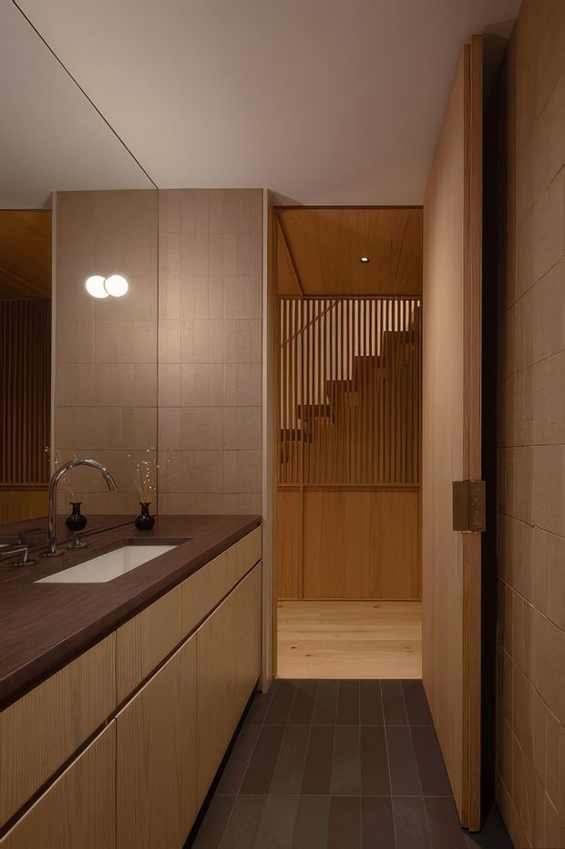 Warm, refined bathroom interior with a long timber-veneered vanity, integrated white basin, dark stone benchtop, wall-to-wall mirror, and large beige tiled walls. A timber-lined opening leads to a stairway screened by vertical timber battens, while dark rectangular floor tiles contrast with the pale timber beyond. Soft pendant lighting and concealed illumination create an intimate atmosphere.