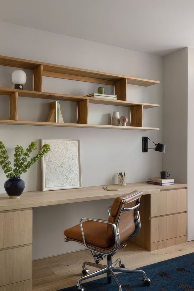 A compact, refined home office with a built-in light timber desk and integrated drawer storage. Open timber shelving spans the wall above the desk, displaying books, ceramics, and decorative objects. A brown leather and chrome swivel office chair sits at the workstation, with a black wall-mounted task lamp, framed artwork, desk accessories, and a plant adding character. The room has light neutral walls, timber flooring, and a calm contemporary aesthetic.