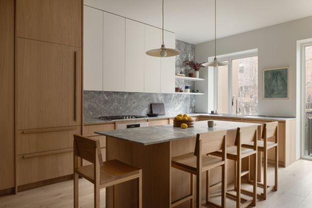 Warm, contemporary kitchen with extensive light-to-medium timber cabinetry, a large central island with integrated seating, white upper cabinets, and a grey veined stone backsplash and benchtop. Pendant lights hang above the island and a dining-height counter extends toward large windows and a glazed door. The room has a calm, refined palette of timber, white, grey, and soft neutral walls, with open shelving and minimal decor.