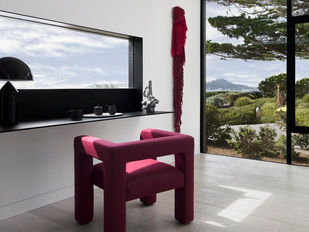 Contemporary interior vignette featuring a sculptural deep burgundy upholstered armchair beside a minimalist built-in black display ledge and large black-framed windows. The light-toned timber floor and white walls create a bright, refined setting, with a landscaped garden and distant coastal landscape visible outside.