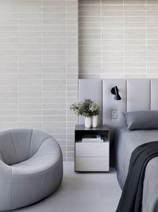 A calm, contemporary bedroom featuring a grey upholstered bed with padded vertical headboard panels, a matching rounded lounge chair, dark bedside table, black wall-mounted reading light, and soft neutral bedding. The room has a pale brick feature wall and light timber flooring, with minimal decorative styling.