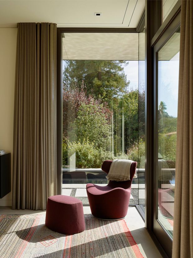 A refined sitting area framed by full-height glazing, with a burgundy sculptural lounge chair and matching ottoman on a patterned rug. Floor-to-ceiling curtains, a ceiling-mounted blind, dark window frames, and abundant garden views create a warm, sunlit indoor-outdoor connection.
