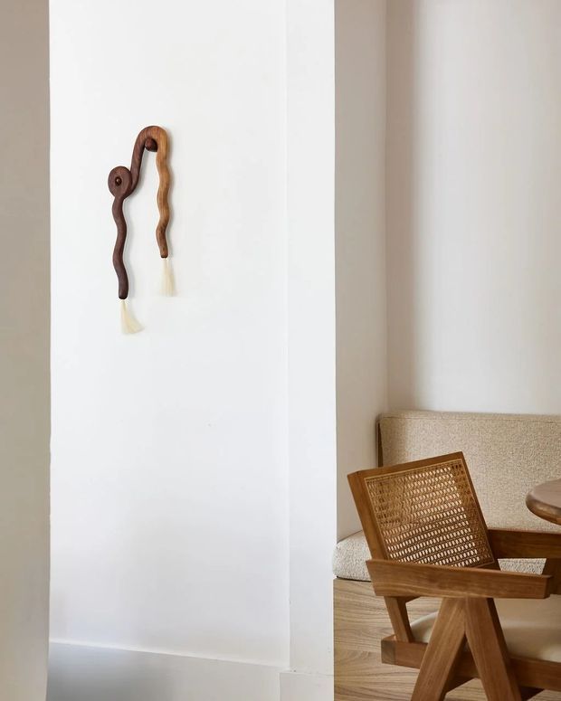 A softly lit minimalist interior vignette featuring a white plaster wall with a small sculptural wooden wall hanging, opening into a warm seating area. The seating area includes a woven cane lounge chair with a timber frame, a beige upholstered sofa, timber flooring, and part of a round wooden table. The palette is neutral and warm with natural wood and woven textures.