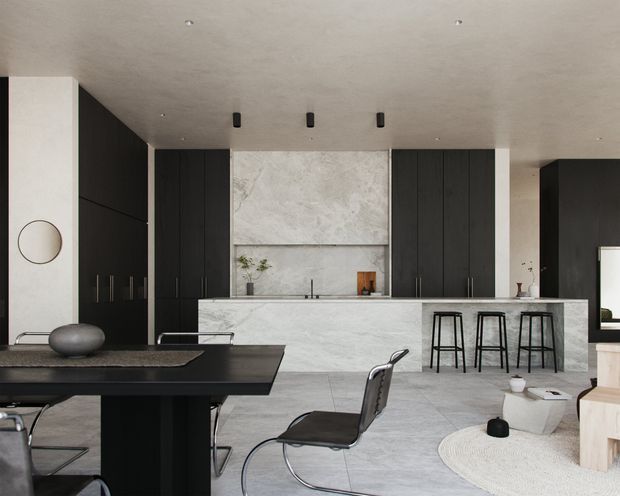 Contemporary open-plan kitchen and dining space with a dramatic black-and-white palette. A large dark dining table and sculptural chairs sit in the foreground, while the rear kitchen features tall matte-black cabinetry, a pale veined stone feature wall and island, integrated appliances, and three bar stools. The space has a smooth pale ceiling with recessed and small black pendant-style spotlights, pale stone flooring, and minimal decorative styling.