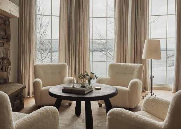Warm, refined living room arranged around a dark rounded coffee table, with several cream boucle armchairs, tall beige curtains, a neutral rug, and a floor lamp. Large divided-pane windows provide expansive snowy outdoor views, while a stone fireplace edge is visible at left.