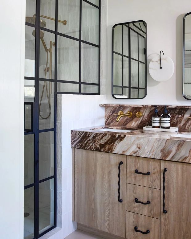 A refined bathroom interior featuring a glass-enclosed shower with black metal grid framing, pale vertical wall tiles, a timber vanity with ornate dark hardware, and a dramatic brown-and-white veined stone benchtop and splashback. Gold-toned wall-mounted tapware, arched grid-framed mirrors, and a round wall light add decorative contrast.
