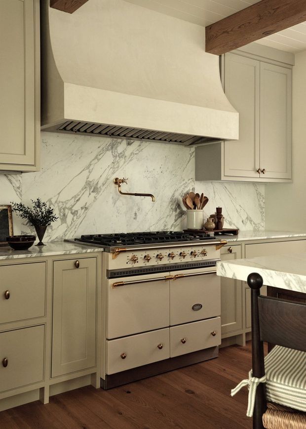 A warmly styled traditional kitchen featuring a large freestanding cream range cooker beneath a sculptural plaster rangehood, framed by pale sage-grey shaker cabinetry. The kitchen has a dramatic white marble backsplash and benchtops with grey veining, brass-toned hardware and tapware, exposed dark timber ceiling beams, and warm timber flooring. A marble-topped island and upholstered dining chair are visible in the foreground.