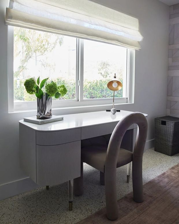 A refined home office or study nook positioned beneath large windows, featuring a sculptural fluted grey desk with rounded corners, a curved upholstered chair, a small gold-and-black desk lamp, a vase of green foliage, and a woven storage basket. The space has soft neutral walls, white window frames, a pale terrazzo-style floor partially covered by a muted rug, and abundant natural light.