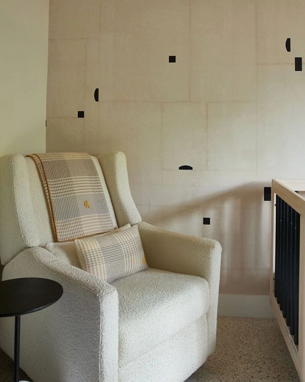A softly lit interior corner featuring a cream boucle upholstered armchair with patterned cushions, a small round black side table, and a pale terrazzo floor. The chair sits against a warm off-white tiled or panelled wall punctuated by small black geometric accents, with a timber-and-black metal stair balustrade visible at right.
