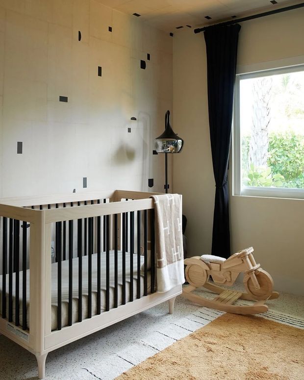 A softly lit nursery with a light timber cot featuring black vertical spindles, a plush neutral rug, a wooden rocking toy, and a black pendant floor lamp. The room has pale walls with sparse dark rectangular accents, a large window with black curtains, and a calm contemporary aesthetic.