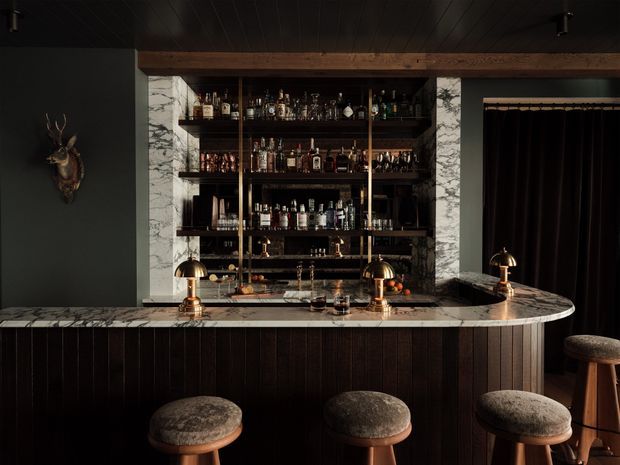 A moody, luxurious home bar with a dark timber-clad counter, rounded marble benchtop, brass dome lamps and brass fixtures. Open shelving behind the counter displays bottles and glassware, framed by dramatic veined marble panels. Upholstered bar stools sit in front, with dark walls, ceiling and curtains creating an intimate atmosphere.