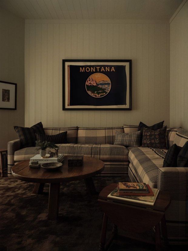 A cozy, dimly lit living room with a large plaid L-shaped sectional sofa, dark patterned cushions, a round wooden coffee table, a wooden side table with books, and a dark area rug. The walls and ceiling are clad in vertical cream-painted timber panelling, with framed vintage-style artwork above the sofa.