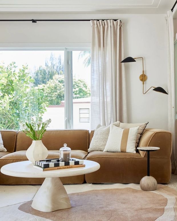 Bright, warm living room with a low caramel-brown leather sectional sofa, textured neutral cushions, a sculptural white coffee table, small round side table, and a large abstract neutral rug. Full-height windows with sheer cream curtains bring in abundant daylight and greenery views. A black-and-brass geometric wall sconce provides a graphic accent, while decorative ceiling moulding adds a refined architectural detail.