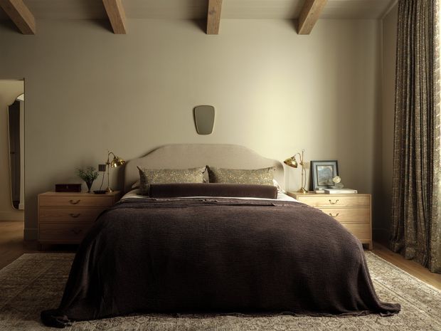 Warm, refined bedroom with a centered upholstered bed dressed in layered neutral and dark bedding, flanked by mismatched wooden bedside tables with lamps and decor. The room features exposed timber ceiling beams, taupe walls, a textured area rug, and full-height patterned curtains.