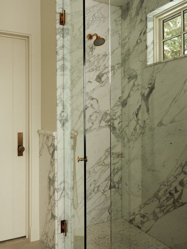 A refined bathroom shower enclosure with floor-to-ceiling veined marble surfaces, a clear glass shower screen and door, and warm brass shower hardware. A small multi-pane window provides natural light, while a white paneled door and marble threshold complete the traditional, luxurious setting.