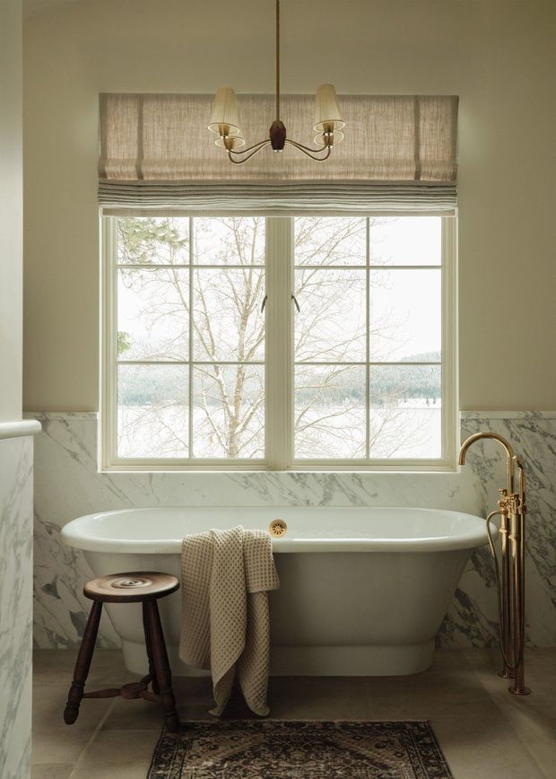 A warm, traditionally styled bathroom centred on a freestanding white clawfoot-style bathtub beneath a large divided window. The room features marble-look wall wainscoting, a Roman shade, aged brass plumbing fixtures, a small dark wooden stool, patterned rug, and a vintage-inspired pendant light. Natural daylight and muted neutral tones create a refined, inviting atmosphere.