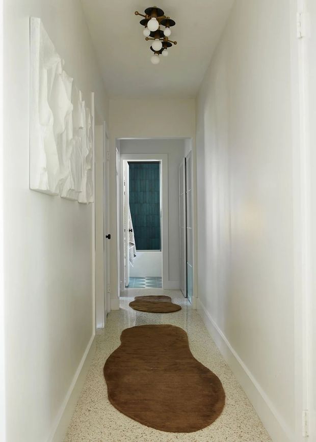 A narrow, bright residential hallway with white walls and trim, a polished terrazzo floor, and organic brown hide rugs running along the corridor. A sculptural mid-century-style pendant light hangs centrally from the ceiling. White doors open toward a small bathroom or utility space at the far end, where teal wall tiling and patterned floor tiles are visible. Abstract white wall art decorates the left wall.