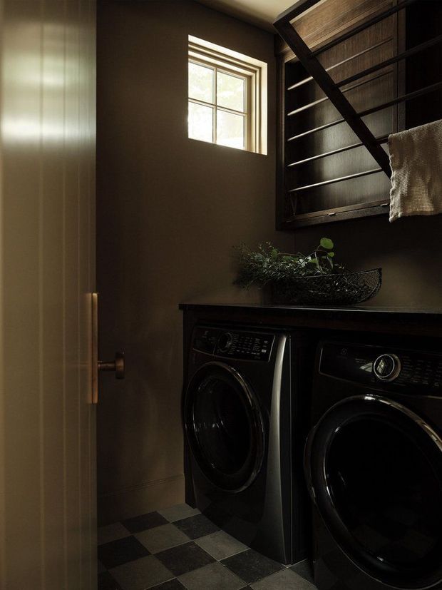 A compact, moody laundry room with dark brown walls, black front-loading washer and dryer beneath a dark countertop, and a black checkerboard tile floor. A small window provides natural light, while a wall-mounted drying rack and hanging towel add practical utility. A black wire basket with greenery sits on the counter.