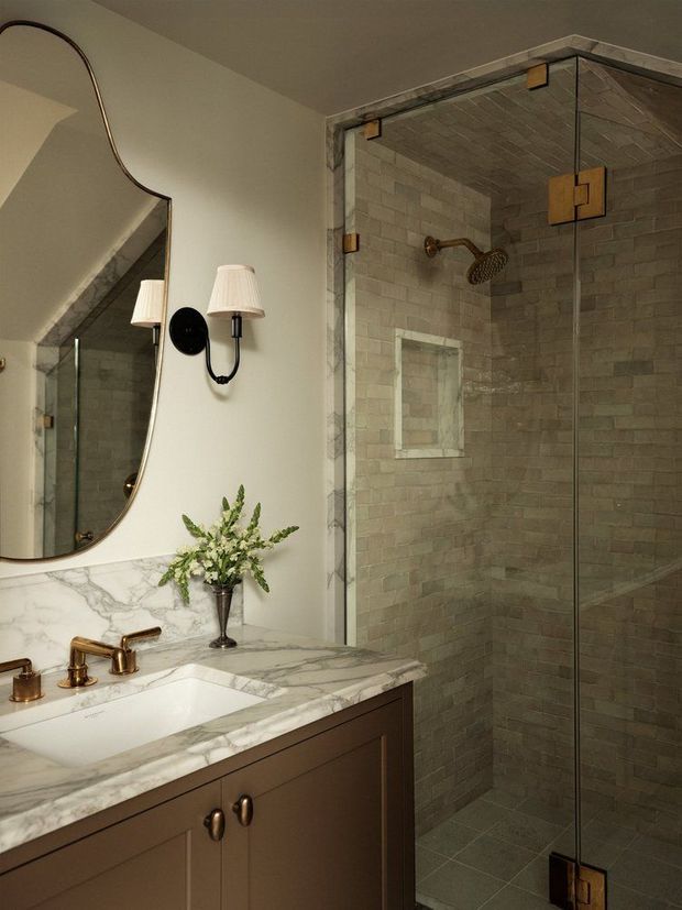 Elegant bathroom featuring a marble vanity countertop with integrated white sink, dark wood cabinetry, brass tapware, a large sculptural wall mirror, decorative wall sconce, and a frameless glass walk-in shower lined with grey stone-look tiles and brass hardware.