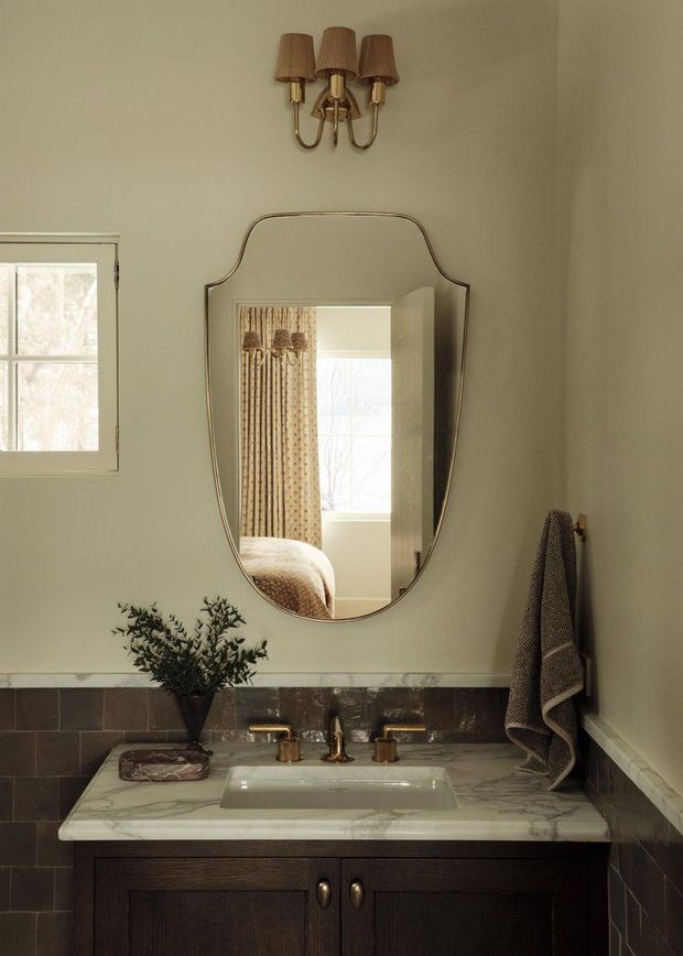 A warm, traditionally styled bathroom vanity with a dark timber cabinet, white marble countertop, integrated rectangular basin, brass tapware, dark wall tiles, and a large curved vintage-style mirror. A three-light brass wall sconce, small window, and towel contribute to the refined, softly lit atmosphere. The mirror reflects an adjacent bedroom area with curtains and a bed.