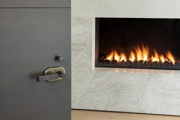 Close-up interior view of a partially open grey timber door with dark metal hardware beside a recessed linear gas fireplace framed in pale veined stone. The image focuses on refined built-in finishes and the warm fire feature.