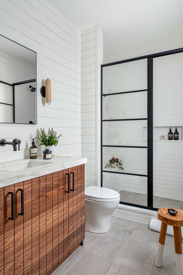 Bright contemporary bathroom featuring white horizontal wall panelling and white subway-tile shower walls, a timber vanity with black handles, a white stone-look benchtop, black wall-mounted tapware, and a black-framed glass shower enclosure. A toilet, mirror, wall sconce, and small timber stool complete the room.
