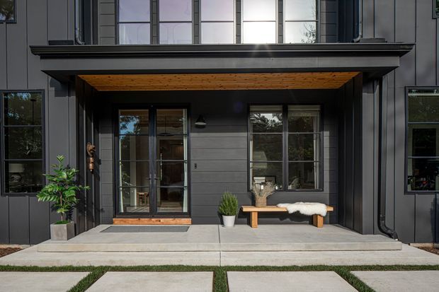 Front exterior entrance of a contemporary multi-storey home, featuring dark vertical cladding, large black-framed windows, a projecting entry canopy with a warm timber soffit, glazed double doors, concrete paving, and a simple bench with potted plants.