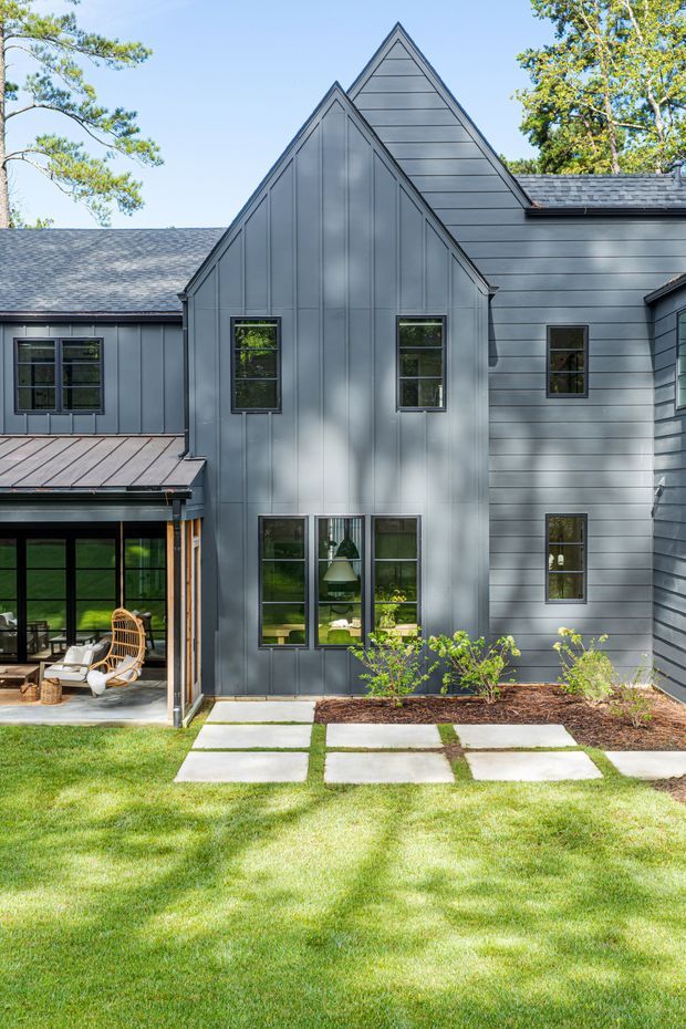 Exterior view of a contemporary multi-volume house with steep front gables, dark grey horizontal siding, charcoal shingle roofs, and large black-framed windows. A covered outdoor seating area opens onto a landscaped lawn with concrete stepping slabs and planted garden beds.