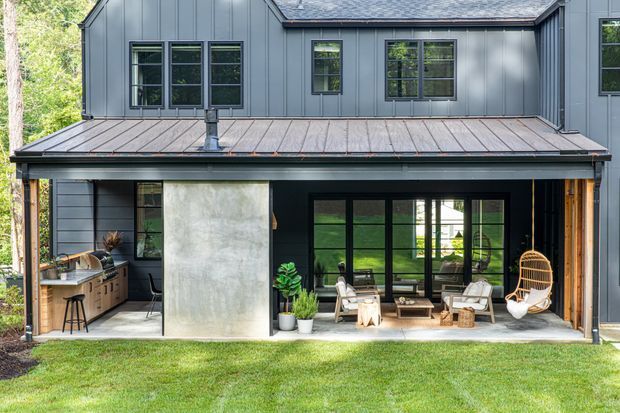 Two-storey contemporary house exterior with dark vertical metal cladding, a standing-seam metal roof, and black-framed windows and doors. The ground floor opens onto a covered outdoor living area with a compact outdoor kitchen at left, concrete feature wall, lounge seating, and a hanging chair. A lawn and wooded setting surround the home.