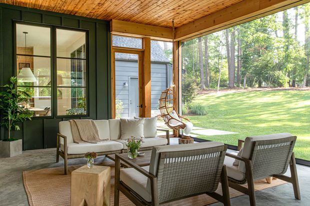 A bright, covered outdoor lounge or sunroom with large glazed openings, a warm timber-lined ceiling, dark green wall panelling, and a relaxed seating arrangement facing a landscaped lawn and wooded setting. The space includes a sofa, lounge chairs, a coffee table, plants, and a hanging chair, creating an airy indoor-outdoor connection.
