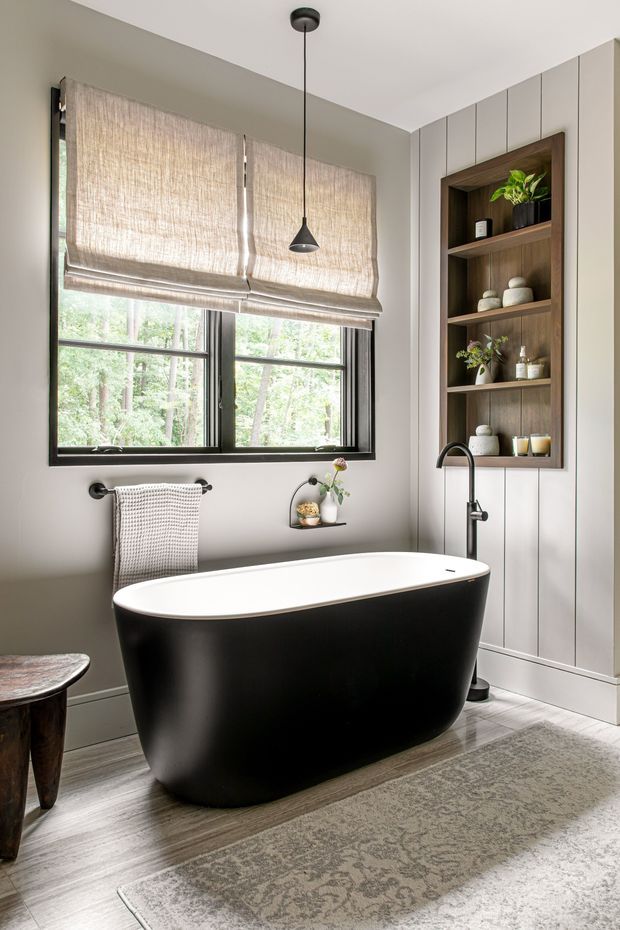 A refined bathroom featuring a freestanding matte-black bathtub with white interior, black-framed windows, a black wall-mounted basin tap, and a built-in timber shelving niche. Soft Roman blinds, vertical light wall panelling, pale timber-look flooring, and a muted patterned rug create a warm, contemporary interior with a view to greenery outside.