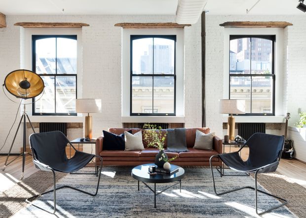 Bright loft-style living room with exposed white-painted brick walls, three tall black-framed industrial windows, exposed timber lintels, and an exposed white duct. The room is furnished with a cognac leather sofa, two black sling lounge chairs, a round black coffee table, table lamps, and a large neutral patterned rug. Strong daylight and city views create an airy, refined urban atmosphere.