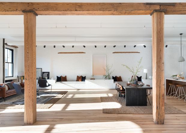Bright open-plan interior featuring a living and dining area with white-painted brick walls, exposed timber posts and beams, pale timber flooring, and large windows. A white built-in sofa or banquette spans the rear wall, while a dark dining table and kitchen or bar joinery occupy the right side. The space combines rustic structural timber with clean contemporary furnishings and abundant natural light.