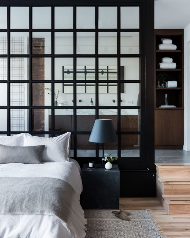 A refined bedroom with a partially visible bed dressed in white and grey linens, a dark bedside table and lamp, and a large black-framed industrial-style glazed partition. Through the partition is a compact bathroom with a marble-topped vanity, dark cabinetry, and black fixtures. Built-in dark timber shelving displays folded towels, while light timber stairs and a patterned rug add warmth. The palette is monochromatic with timber and exposed brick accents.