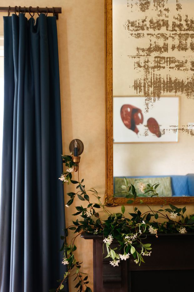 A warmly styled interior vignette featuring floor-length deep navy curtains on a dark timber rod, a textured beige wall, an ornate timber-framed mirror, framed abstract artwork, a wall-mounted candle sconce, and trailing greenery arranged across a dark timber mantel or console. The mirror reflects blue seating and additional greenery.