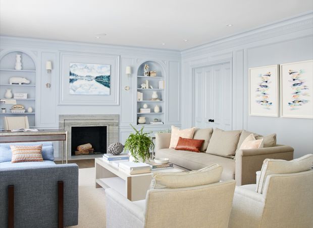 Bright, refined living room with pale blue-gray paneled walls, ornate trim, built-in arched display shelving, a classic fireplace with stone surround, and elegant neutral upholstered seating arranged around a white coffee table. Coastal artwork, soft furnishings, and natural greenery add a relaxed yet polished character.
