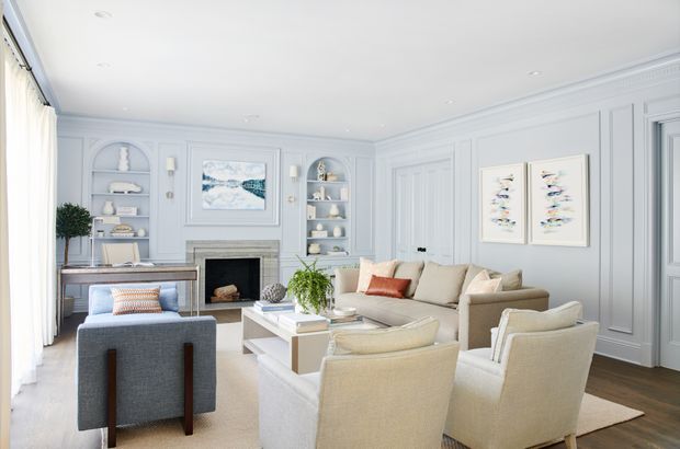 A bright, refined living room with pale blue-grey paneled walls, detailed decorative mouldings, built-in display shelving, and a traditional fireplace. The room includes a neutral sectional sofa, upholstered armchairs, a glass coffee table, a console desk, soft cream curtains, and coastal-toned artwork. Natural light and a restrained palette create an airy, elegant atmosphere.