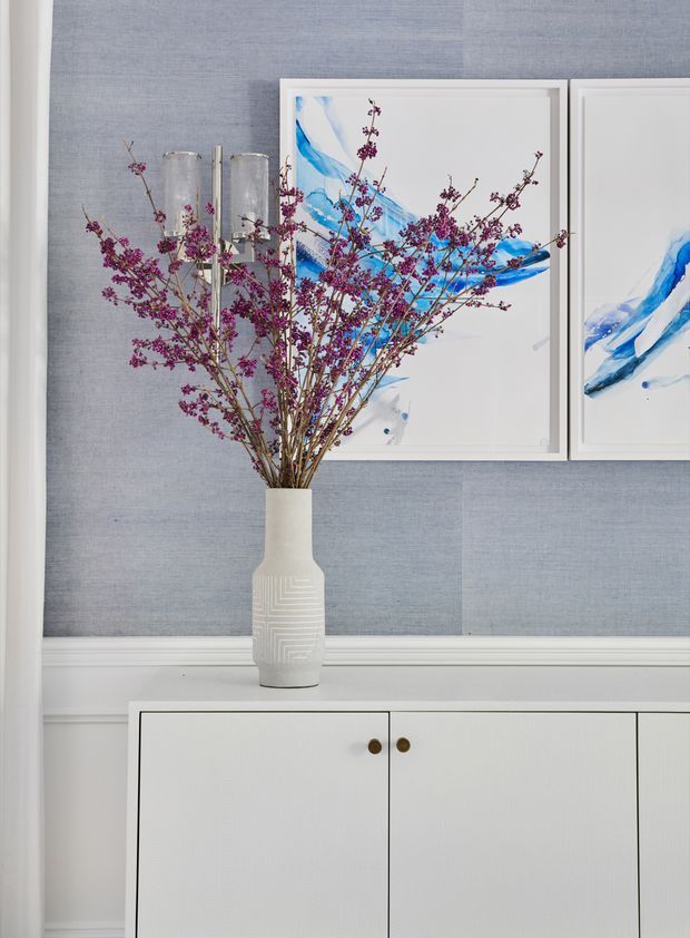 A refined interior vignette featuring a white built-in sideboard with small round brass-toned knobs, set against pale blue textured wallcovering and white trim. A sculptural white vase with flowering branches stands in front of framed blue-and-white abstract artwork, accompanied by tall glass candleholders.