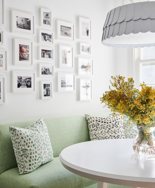 Bright, airy sitting area with a pale mint-green upholstered banquette wrapping around a round white table. Patterned green and black-and-white cushions add contrast, while a gallery wall of framed photographs decorates the white wall. A large scalloped pendant light hangs near a bright window, beside a clear vase of yellow flowering branches. The overall look is playful, fresh, and refined.