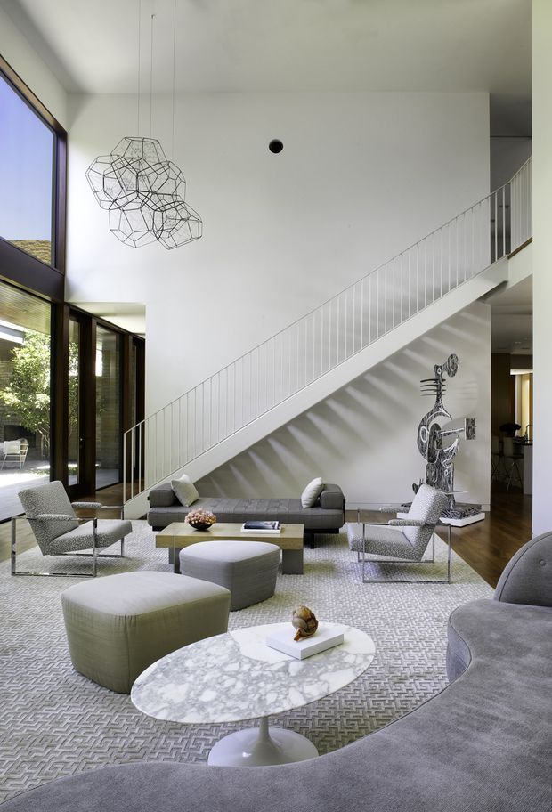 A spacious, double-height contemporary living room with a dramatic white staircase and slender white balustrade. Floor-to-ceiling glazing and dark timber-framed doors open toward an outdoor courtyard. The room is furnished with multiple grey upholstered lounge chairs, sofas, ottomans, and white marble oval pedestal coffee tables over a patterned neutral rug. A sculptural wire pendant light hangs above the seating area, while a large sculptural artwork sits beneath the stairs. The space has a refined, gallery-like feel with abundant natural light.