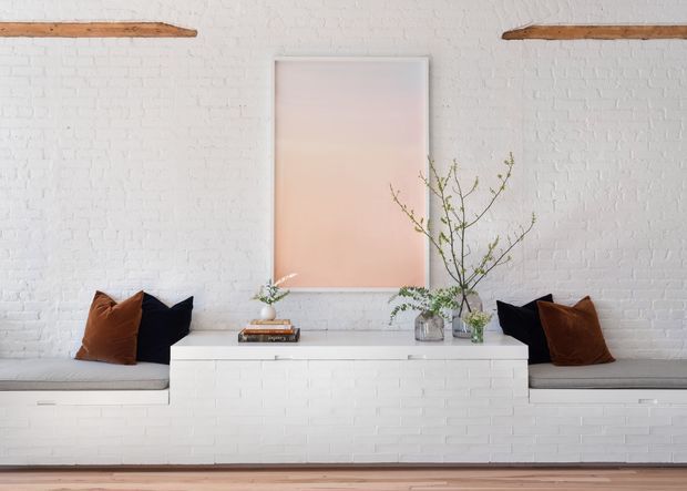 Bright, minimalist interior with white painted brick walls and a long built-in white brick bench or console arrangement. Grey upholstered bench seating runs along both sides with black and warm brown cushions. A pale pink abstract artwork is centered on the wall, while simple branches in glass vessels add an organic accent. Exposed timber ceiling beams and warm timber flooring introduce contrast.