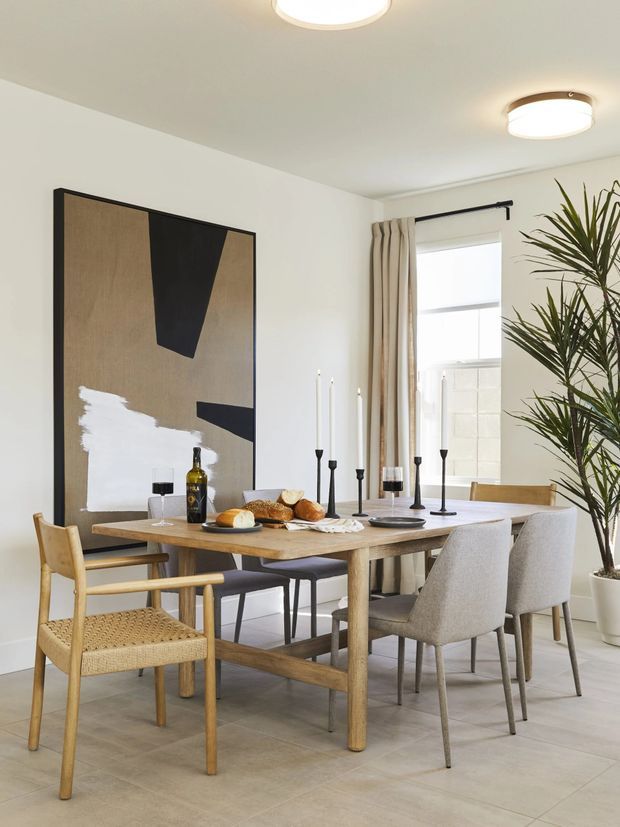 Bright, refined dining room with a large timber dining table surrounded by upholstered dining chairs and a woven armchair. The room features pale tiled flooring, white walls and ceiling, full-height curtains, a large abstract artwork, simple contemporary ceiling lights, black candleholders, and a tall indoor plant beside the window.