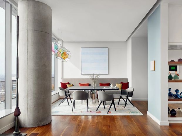 A refined open-plan dining and living area in a high-rise apartment, featuring floor-to-ceiling glazing, a large cylindrical concrete structural column, polished timber flooring, a contemporary dining table with sculptural chairs, and a built-in display shelving unit. The neutral interior is enlivened with colourful abstract artwork and cushions, while city views are visible through the windows.