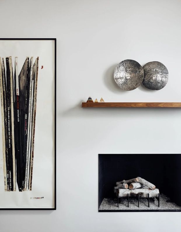 A close-up view of a light grey painted interior wall featuring curated wall art and a narrow floating timber shelf. The shelf displays small geometric objects and two overlapping circular metallic sculptures. A large framed abstract artwork hangs at left, while a dark recessed fireplace or niche at lower right contains a small sculptural arrangement. The composition is refined and minimalist with strong monochrome, timber, and metallic accents.