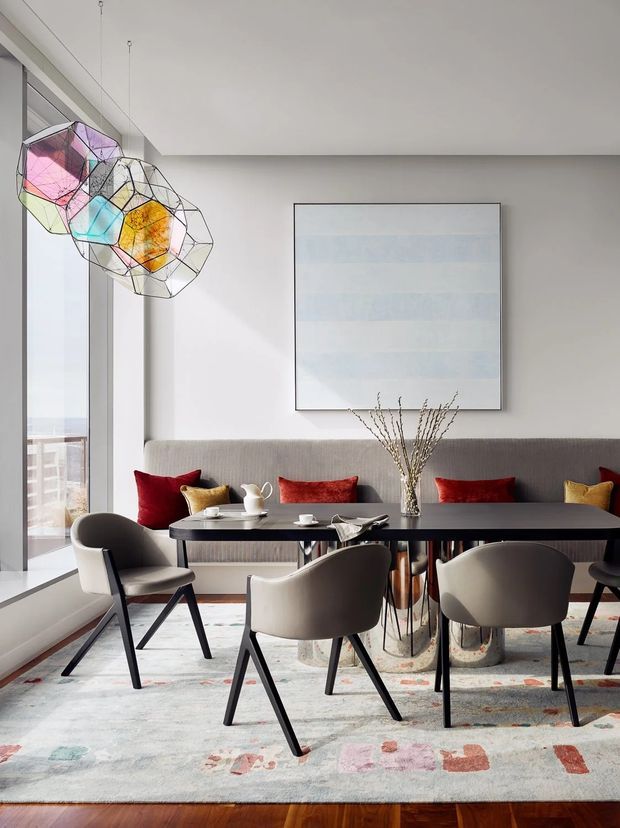 A bright, refined dining area with a long dark dining table surrounded by upholstered modern chairs. A built-in banquette lines the back wall with deep red and mustard cushions, while a large pale abstract artwork hangs above it. Floor-to-ceiling glazing opens to a balcony or exterior view, and sculptural geometric pendant lights add a colourful feature above the table. A patterned area rug and timber flooring complete the space.