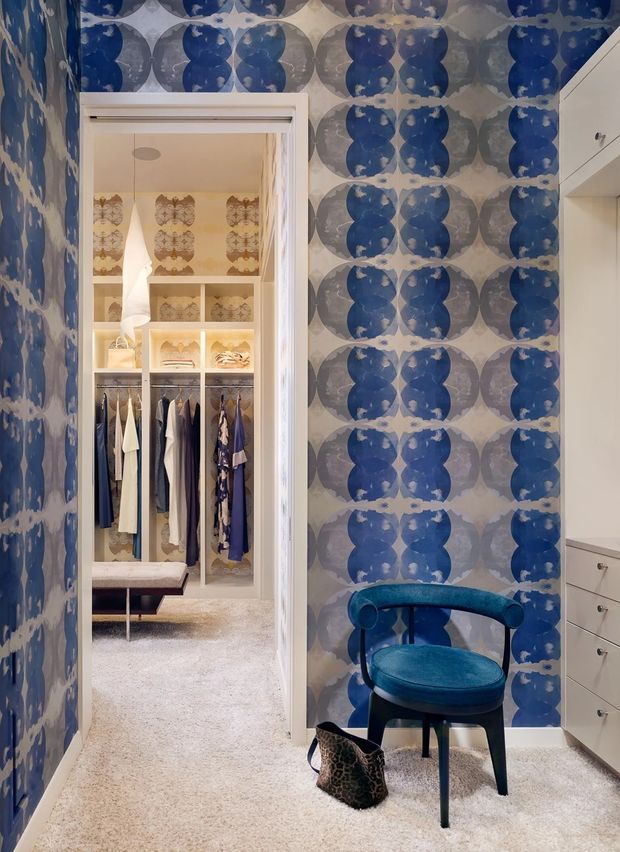 A colourful walk-in wardrobe or dressing area with dramatic blue and grey patterned wallpaper, white built-in drawers and cabinetry, and a teal upholstered chair. Through the open doorway, a carpeted dressing room with hanging clothes, shelving, and a bench is visible. The space has a playful, highly decorative and refined interior.