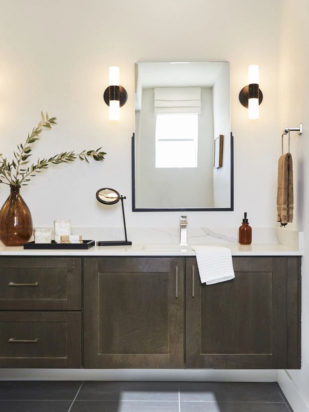 A refined contemporary bathroom vanity with dark wood cabinetry, a white stone countertop, integrated basin and chrome tapware. A large black-framed mirrored cabinet is flanked by cylindrical wall sconces, with a small window reflected in the mirror. Dark tiled flooring, a brown glass vase with foliage, towel rail and amber soap dispenser complete the space.